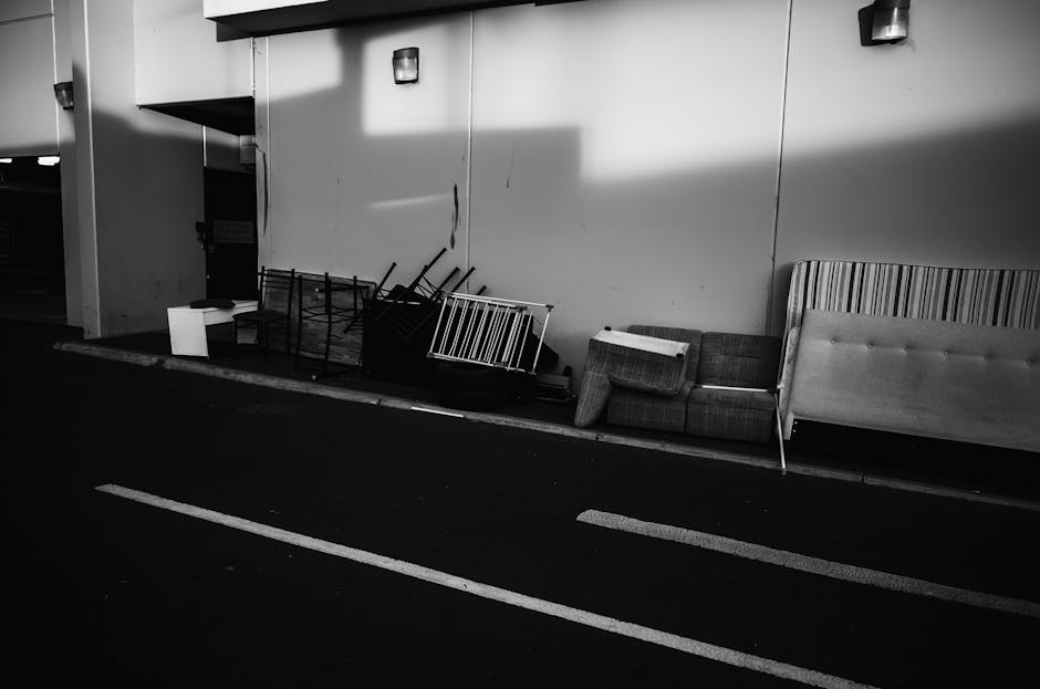 Black-and-white image showing a section of a building’s interior or possibly a parking area with a plain wall on the right side, partially illuminated by overhead lighting. In front of the wall, there are discarded or stored furniture items and packaging materials, including a small armchair, a larger upholstered sofa or bed frame, and a striped panel or headboard. Several disassembled or stacked metal bed frames, some wrapped in plastic or secured with straps, are leaning against the wall. There are also cardboard boxes and packing materials scattered around, indicating a recent or ongoing furniture removal process. The floor appears to be paved, with some white lines marking parking spaces or pathways. The scene suggests a furniture collection or disposal area, which could be part of home relocation services offered by Man with Van Cowley as they carry out loading, packing, and furniture transport for removals or clearance projects.