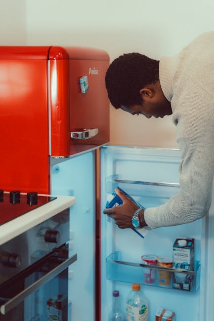 A man with dark skin and short curly hair is leaning over to open an empty fridge in a kitchen, holding a smartphone in his right hand. The fridge has a bright blue interior and is positioned next to a red kitchen appliance, possibly a microwave or small oven. The scene is illuminated with natural and artificial light, and the man is dressed in a light grey, long-sleeved sweater. To the left, part of a white countertop with a gas stove featuring black burners and control knobs is visible. This setting suggests the process of organizing or preparing for a home relocation, with a focus on packing or clearing space as part of a furniture transport or moving service. The image is associated with professional removals provided by Man with Van Cowley, supporting house removals and packing solutions, especially relevant for services like bulky item pickup and home relocation in OX4.