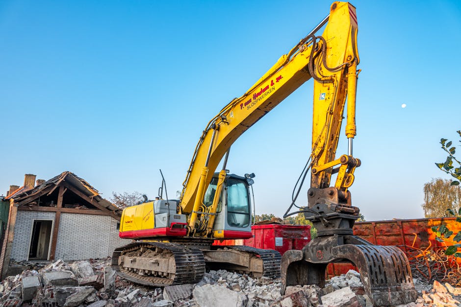 A yellow excavator equipped with a hydraulic arm and claw attachment is positioned on a pile of broken bricks and rubble outside a partially demolished building. The excavator's cab features tinted wi