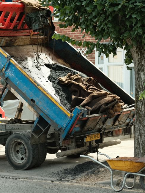 A yellow excavator equipped with a hydraulic arm and claw attachment is positioned on a pile of broken bricks and rubble outside a partially demolished building. The excavator's cab features tinted wi