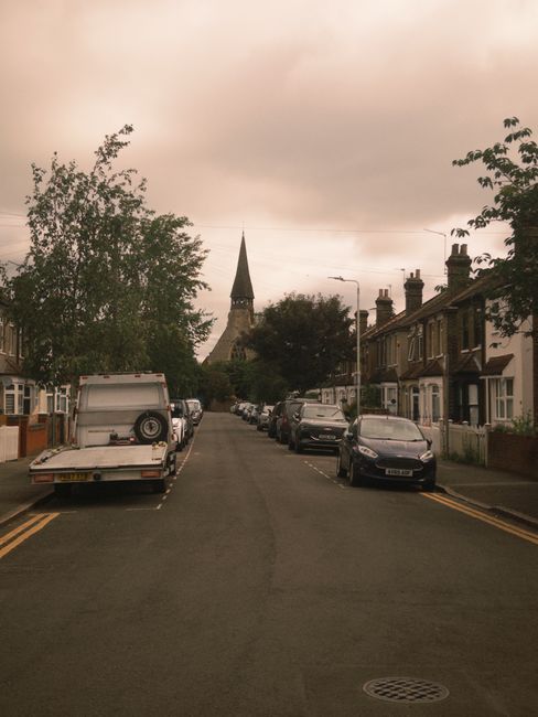 A narrow residential driveway during daytime, with several parked cars lined up along the left side, including dark-colored hatchbacks and sedans, partially covered in shadows from overhanging trees. 