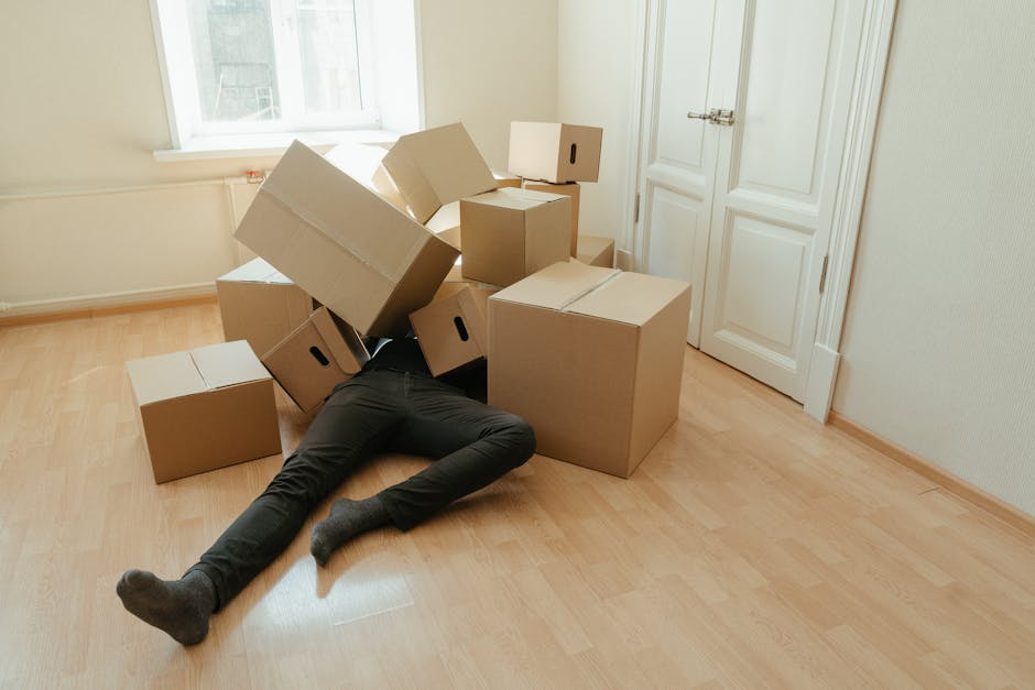 A person is lying face down on a light wooden floor in a room with cream walls and a large window that allows natural light to illuminate the space. The individual is partially concealed beneath a stack of cardboard boxes of various sizes, some of which are open and others closed, with the boxes arranged haphazardly around their body. Several boxes are positioned near a white door with a metallic handle and hinges. The boxes appear to be packed with household items for a home relocation, with some boxes stacked on each other. There are no other visible furnishings or items in the room. The scene depicts the process of loading or packing during furniture transport or packing and moving activities, typical of professional removals, as carried out by companies such as Man with Van Cowley.