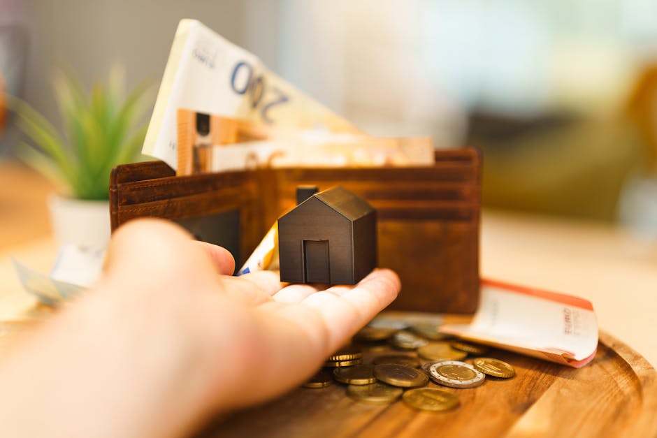 A close-up view of a person's hand placing a small black house-shaped model into a brown leather wallet that contains some 20 and 50-pound currency notes. The wallet rests on a wooden surface alongside some scattered coins and a folded British banknote. In the background, there is a potted plant and additional blurred banknotes, while natural light illuminates the scene, highlighting details relevant to home relocation, packing, and financial planning involved in house removals. The image emphasizes the importance of budgeting and financial awareness when organising a move, as offered by Man with Van Cowley in their removals services.