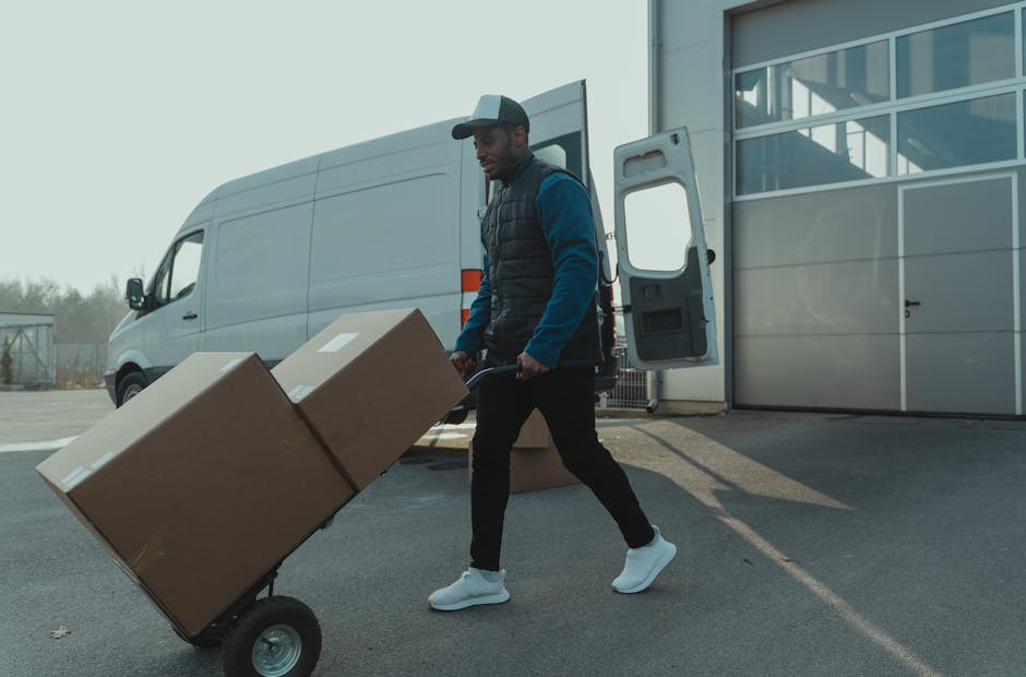 A man wearing a grey cap, blue long-sleeve shirt, and black puffer vest is seen loading two large cardboard boxes onto a hand trolley outside a commercial building during daylight. The boxes are seale