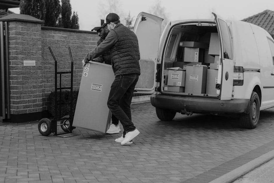 A man wearing a grey cap, blue long-sleeve shirt, and black puffer vest is seen loading two large cardboard boxes onto a hand trolley outside a commercial building during daylight. The boxes are seale