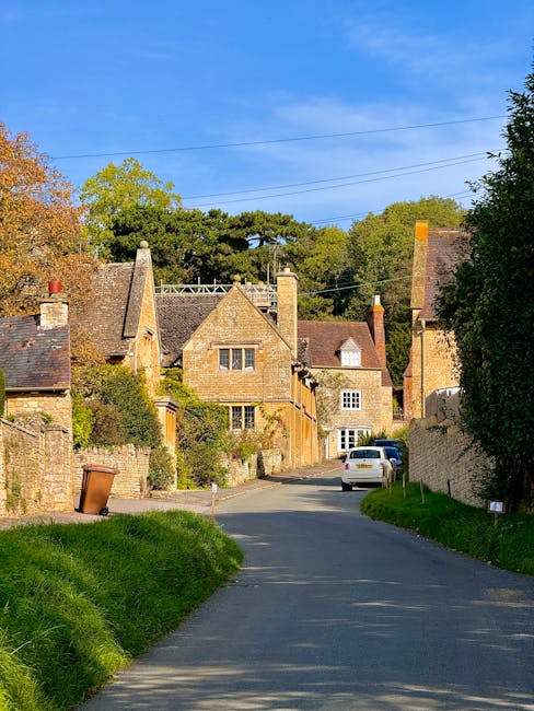A narrow residential street in Iffley Village featuring historic brick houses with pitched roofs, some with small windows and visible chimneys. The street is paved with asphalt and has a slight inclin