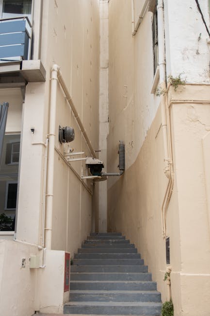A narrow outdoor staircase located between two tall, beige-painted building walls, with six concrete steps leading upward. The walls have visible piping, including white plastic pipes and a black utility box attached to the left side. The steps are slightly worn, and on the left side, part of a modern apartment with blue and grey exterior panels is visible. The area appears to be a back alley or service entrance, with minimal natural light illuminating the space. This setting is consistent with urban home relocation contexts, where careful navigation of tight staircases is often necessary during furniture transport or packing and moving operations, as facilitated by Man with Van Cowley.