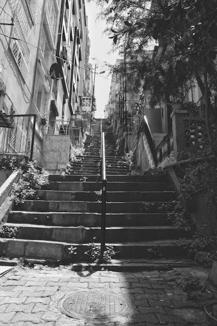 Black and white image showing a steep, narrow staircase made of concrete steps in a Victorian residential area of Cowley, with a metal handrail running up the centre. To the left of the staircase, there are multi-storey houses with brick facades, large windows, and external balconies. The right side features a small terrace with potted plants, a wooden fence, and shrubs. At the bottom of the stairs, cobbled paving leads to a manhole cover in the foreground. The scene is partially shaded by trees, giving a sense of dense urban greenery. The setting appears to be outdoors, with natural light illuminating the scene. The photo captures the challenging access for home relocation or furniture transport in these historic properties, aligning with the services offered by Man with Van Cowley in house removals and moving logistics.