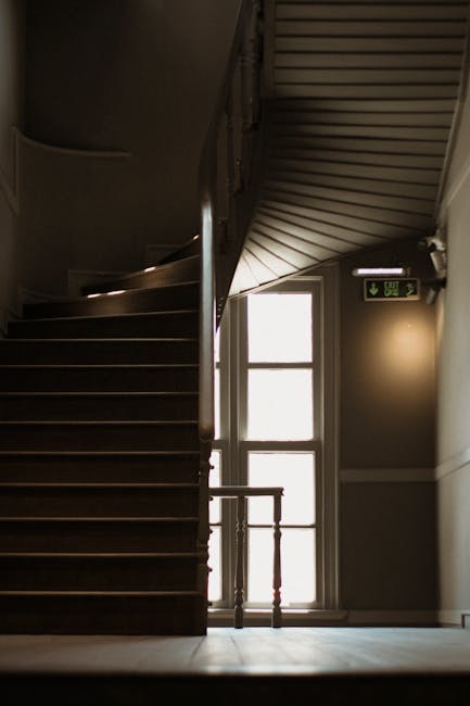 A narrow outdoor staircase located between two tall, beige-painted building walls, with six concrete steps leading upward. The walls have visible piping, including white plastic pipes and a black utility box attached to the left side. The steps are slightly worn, and on the left side, part of a modern apartment with blue and grey exterior panels is visible. The area appears to be a back alley or service entrance, with minimal natural light illuminating the space. This setting is consistent with urban home relocation contexts, where careful navigation of tight staircases is often necessary during furniture transport or packing and moving operations, as facilitated by Man with Van Cowley.