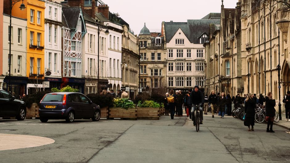 A street scene in Oxford city centre showing a mix of historic and modern buildings lining the narrow pavement. Several parked cars and bicycles are visible along the roadside, with pedestrians walking and gathering near shop entrances. A man is seen riding a bicycle towards the camera in the middle of the street, while others stand in groups, some holding bags or engaged in conversation. Large wooden planter boxes filled with greenery are positioned along the edge of the pavement, separating pedestrians from parked vehicles. The buildings feature a variety of architectural styles, including traditional stone facades and contemporary design elements, with multiple stories and decorative windows. Overcast sky overhead provides diffuse lighting for the scene. This image captures the urban environment where residential and commercial spaces coexist, highlighting the typical setting for home relocation or furniture transport practices, as undertaken by services like Man with Van Cowley.