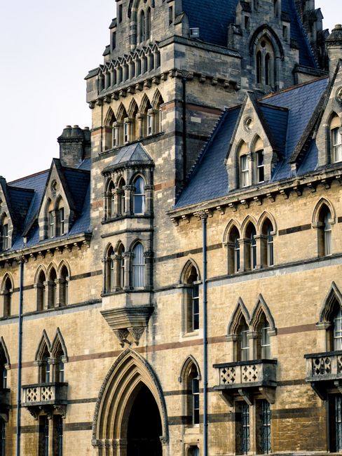 The image shows the exterior of a historic stone building with Gothic architectural features, including pointed arch windows, small decorative balconies with metal railings, and a prominent central tower with arched openings. The building's facade is constructed from light-colored and darker stone blocks, with steeply pitched slate roofs and intricate detailing around the windows and doorways. The surroundings are well-lit by natural daylight, highlighting the textures and details of the stonework. In the context of house removals and relocation services, this image could represent the type of exterior environment where home or office furniture transport may take place, with Man with Van Cowley providing packing and moving expertise near such historic structures, emphasizing careful handling during the loading process and maintaining the building's preservation.