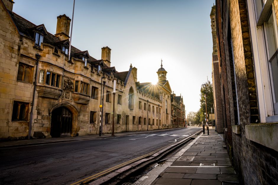 A street scene in Oxford city centre showing a mix of historic and modern buildings lining the narrow pavement. Several parked cars and bicycles are visible along the roadside, with pedestrians walking and gathering near shop entrances. A man is seen riding a bicycle towards the camera in the middle of the street, while others stand in groups, some holding bags or engaged in conversation. Large wooden planter boxes filled with greenery are positioned along the edge of the pavement, separating pedestrians from parked vehicles. The buildings feature a variety of architectural styles, including traditional stone facades and contemporary design elements, with multiple stories and decorative windows. Overcast sky overhead provides diffuse lighting for the scene. This image captures the urban environment where residential and commercial spaces coexist, highlighting the typical setting for home relocation or furniture transport practices, as undertaken by services like Man with Van Cowley.