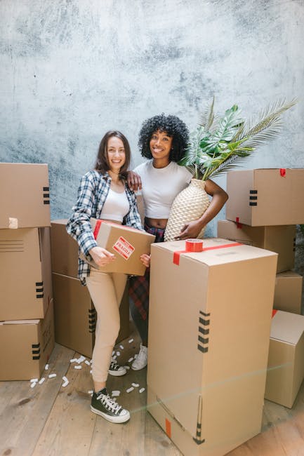 Two women stand inside a room surrounded by stacked cardboard moving boxes, some sealed with red tape. One woman, wearing a plaid shirt and beige pants, holds a small box, while the other woman, dressed in a white top and checkered skirt, holds a large decorative pineapple-shaped vase with green palm leaves. The boxes are positioned on the wooden floor, with additional boxes behind and to the sides, indicating an active packing or home relocation process. The room has a textured, light grey wall and minimal decoration, with soft lighting illuminating the scene. The women are smiling and appear to be engaged in a furniture transport or packing and moving activity, reflecting a professional approach to house removals, as offered by services like Man with Van Cowley.