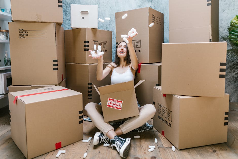 A woman sitting cross-legged on a wooden floor surrounded by numerous large cardboard moving boxes, some sealed with red tape and labeled as fragile, inside a room with pale blue textured walls and green plants. She is wearing a white tank top, beige pants, and black sneakers, and is actively engaged in packing or unpacking, with packing foam pieces falling from her hands into an open box in her lap. The scene depicts the packing process typical of home relocation or furniture transport, with some boxes stacked along the walls and others on the floor, indicating an active moving environment. This visual illustrates the packing and loading aspects of house removals, supported by professional services like those offered by Man with Van Cowley, specialists in quick and efficient relocations.