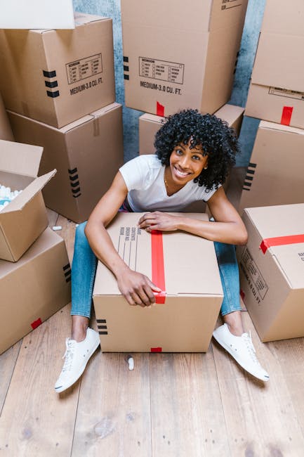 Two women stand inside a room surrounded by stacked cardboard moving boxes, some sealed with red tape. One woman, wearing a plaid shirt and beige pants, holds a small box, while the other woman, dressed in a white top and checkered skirt, holds a large decorative pineapple-shaped vase with green palm leaves. The boxes are positioned on the wooden floor, with additional boxes behind and to the sides, indicating an active packing or home relocation process. The room has a textured, light grey wall and minimal decoration, with soft lighting illuminating the scene. The women are smiling and appear to be engaged in a furniture transport or packing and moving activity, reflecting a professional approach to house removals, as offered by services like Man with Van Cowley.