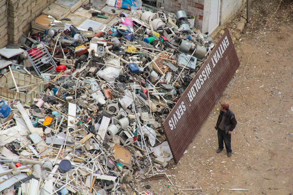 An aerial view of a large disposal site with a significant pile of mixed household waste, including plastic containers, metal and wooden furniture parts, electronic debris, and various discarded domestic items. The waste is spread across a gravel-covered ground, with a large brown sign reading 'FINAL DISPOSAL SERVICES' positioned at the edge of the pile. A man dressed in dark clothing and a brown hat stands nearby, observing the site. The scene is set outdoors, adjacent to a brick building wall, indicating an area designated for the collection and disposal of old or unwanted household items as part of home relocation or house clearance services, illustrating environmental management and waste processing related to removals.