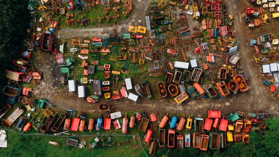 An aerial view of a large outdoor area filled with numerous discarded and abandoned construction and industrial equipment, including metal containers, crates, and various pallets in different sizes and colors. The equipment is scattered across dirt and grass, with some tables, benches, and structural parts stacked or laid out in an irregular pattern. Several vehicles, including a few small trucks and trailers, are parked among the debris, suggesting a site clearance or equipment disposal process. Natural daylight illuminates the scene, highlighting the variety of materials such as wood, metal, and plastic used in the equipment. The area appears to be part of a waste disposal or decommissioned site, which may be relevant for household removals or clearance services such as those offered by Man with Van Cowley. The image captures the complexity of moving, packing, or removing bulky items typically encountered in home relocation or furniture transport scenarios.